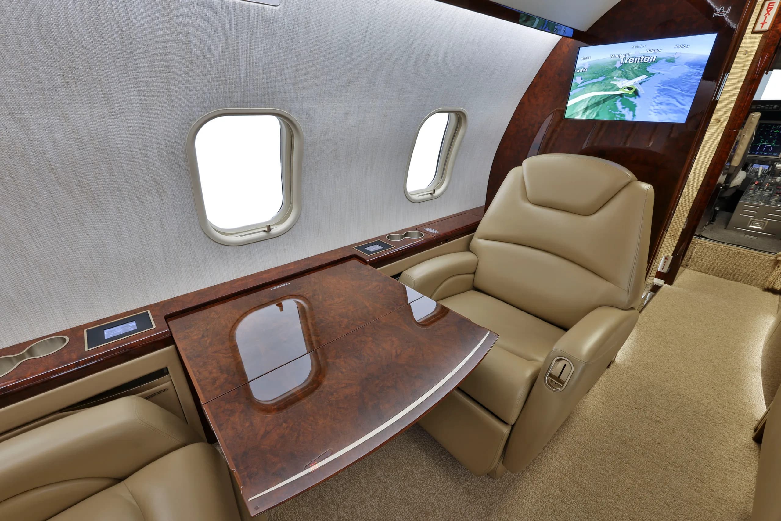 A tan leather seat with a pull-out wooden table and a large display screen showing a map in a business jet cabin.