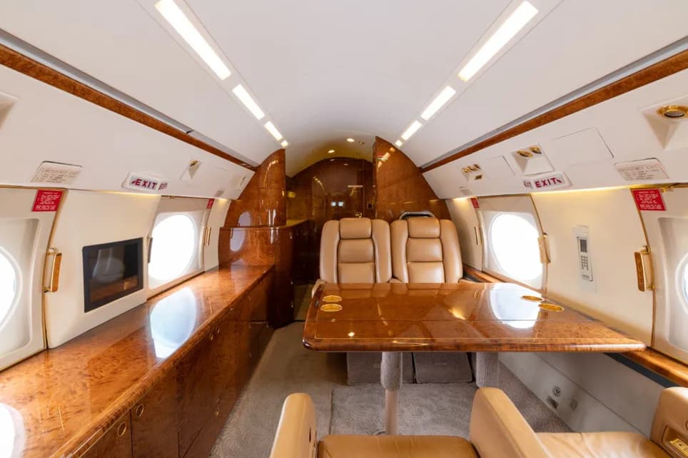 The aft cabin of the business jet with a large polished wood dining table and facing beige leather seats.