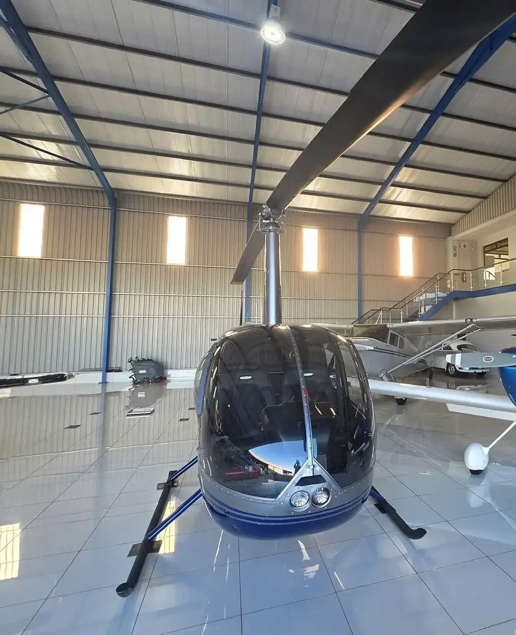 A front view of a silver and blue Robinson R44 Raven II helicopter with its main rotor blade in a hangar.