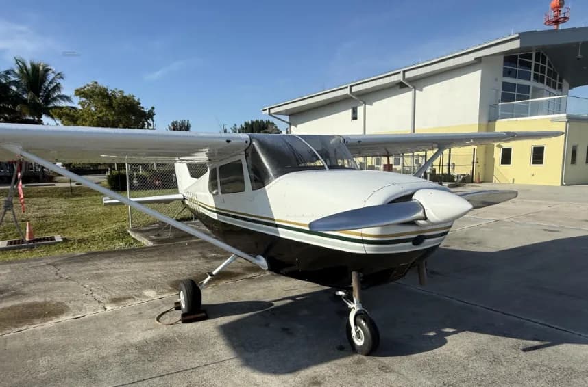 A front-quarter view of a white and black Cessna 172 with gold stripes, parked on an airfield with a building in the background.