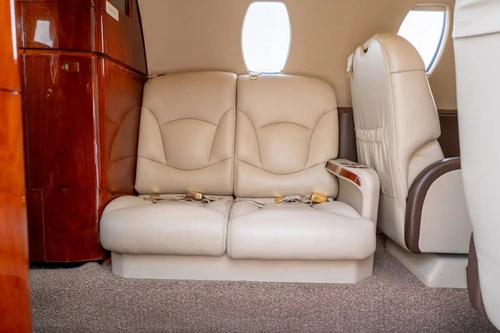A two-person beige leather bench seat with seatbelts in the private jet cabin, next to a wooden cabinet.