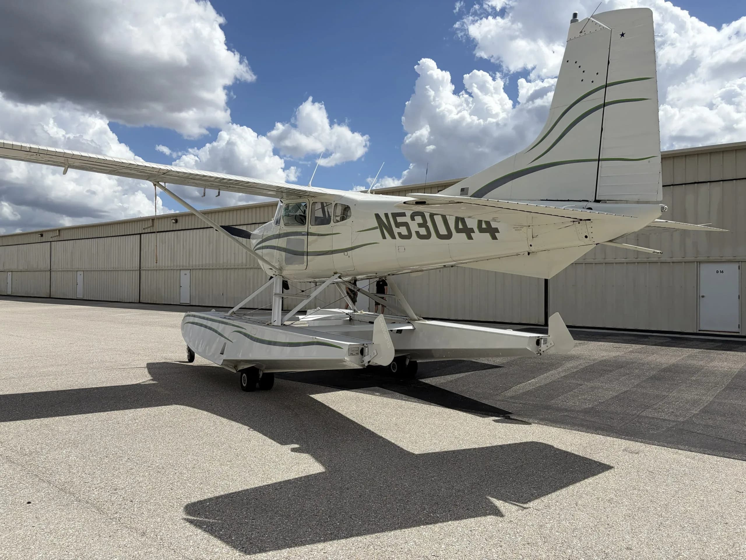 A side view of a white Cessna 180 Skywagon seaplane with green stripes, registration N53044, parked on an asphalt apron.