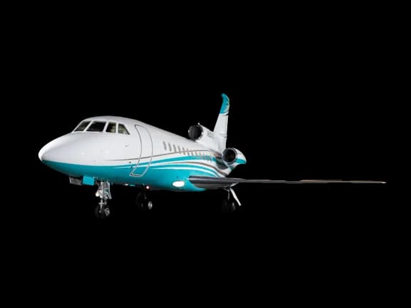 A white and teal business jet, likely a Dassault Falcon, is parked on a dark surface, viewed from the front-left.