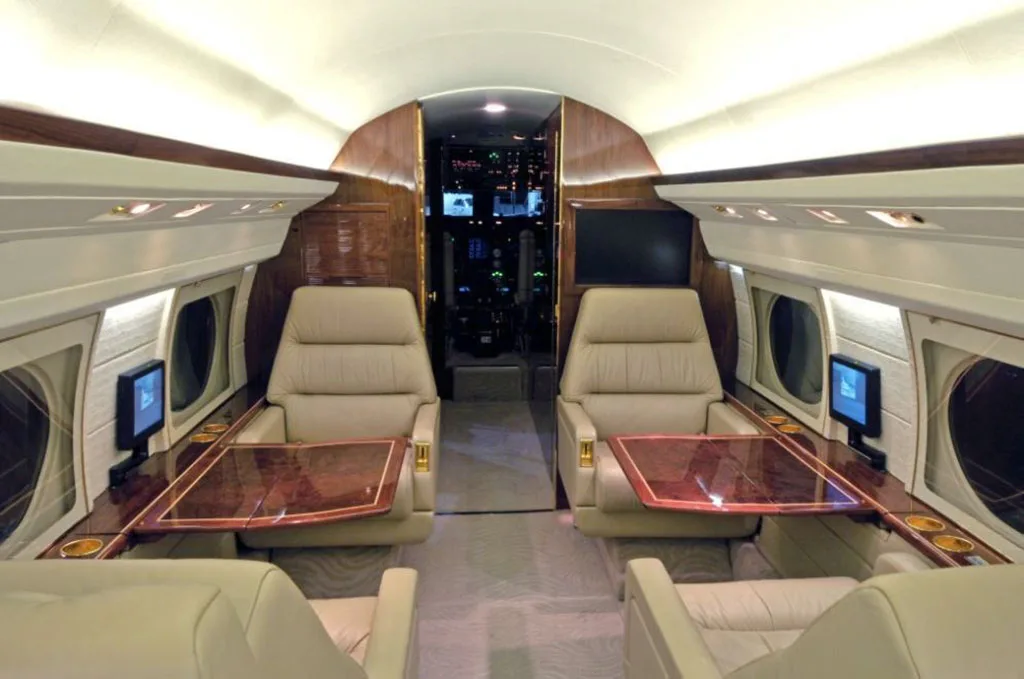 The luxurious interior cabin of a private jet with beige leather seats, wooden tables, and individual screens.