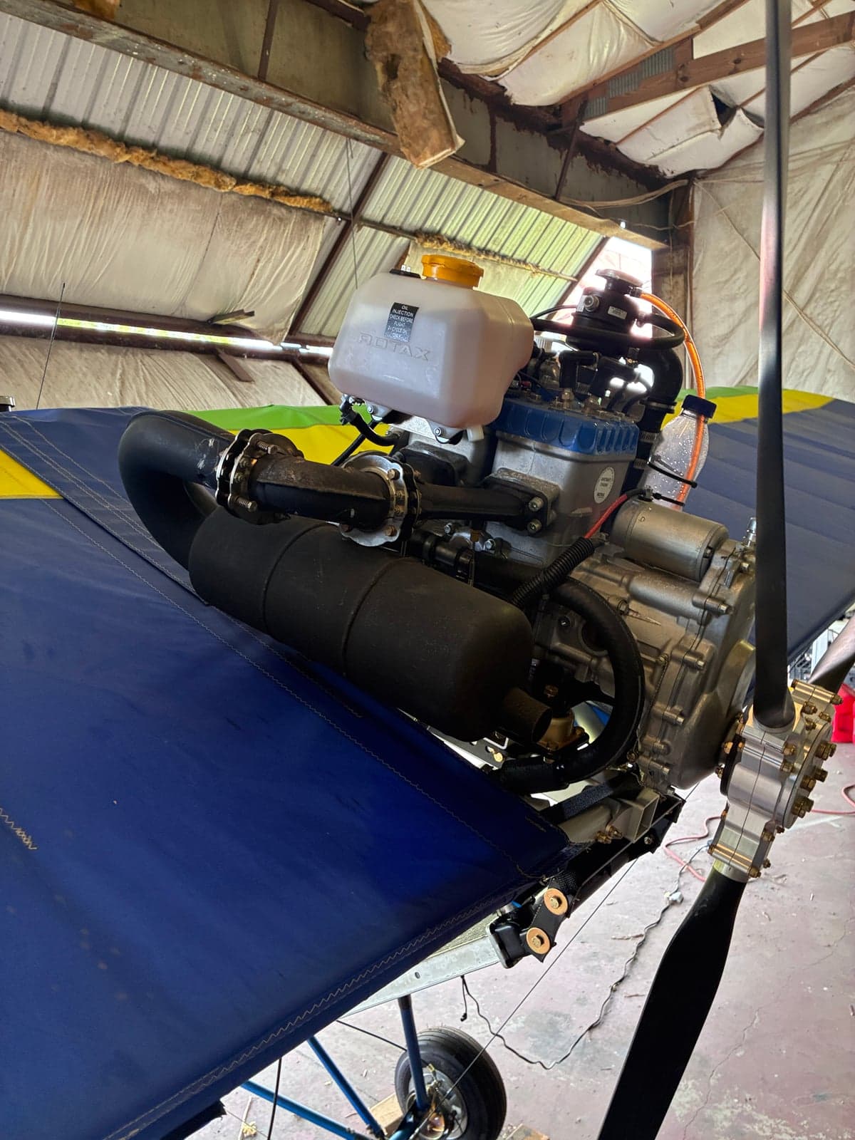 Close-up view of the Rotax engine and propeller mounted on the wing of a blue and yellow ultralight aircraft in a hangar.