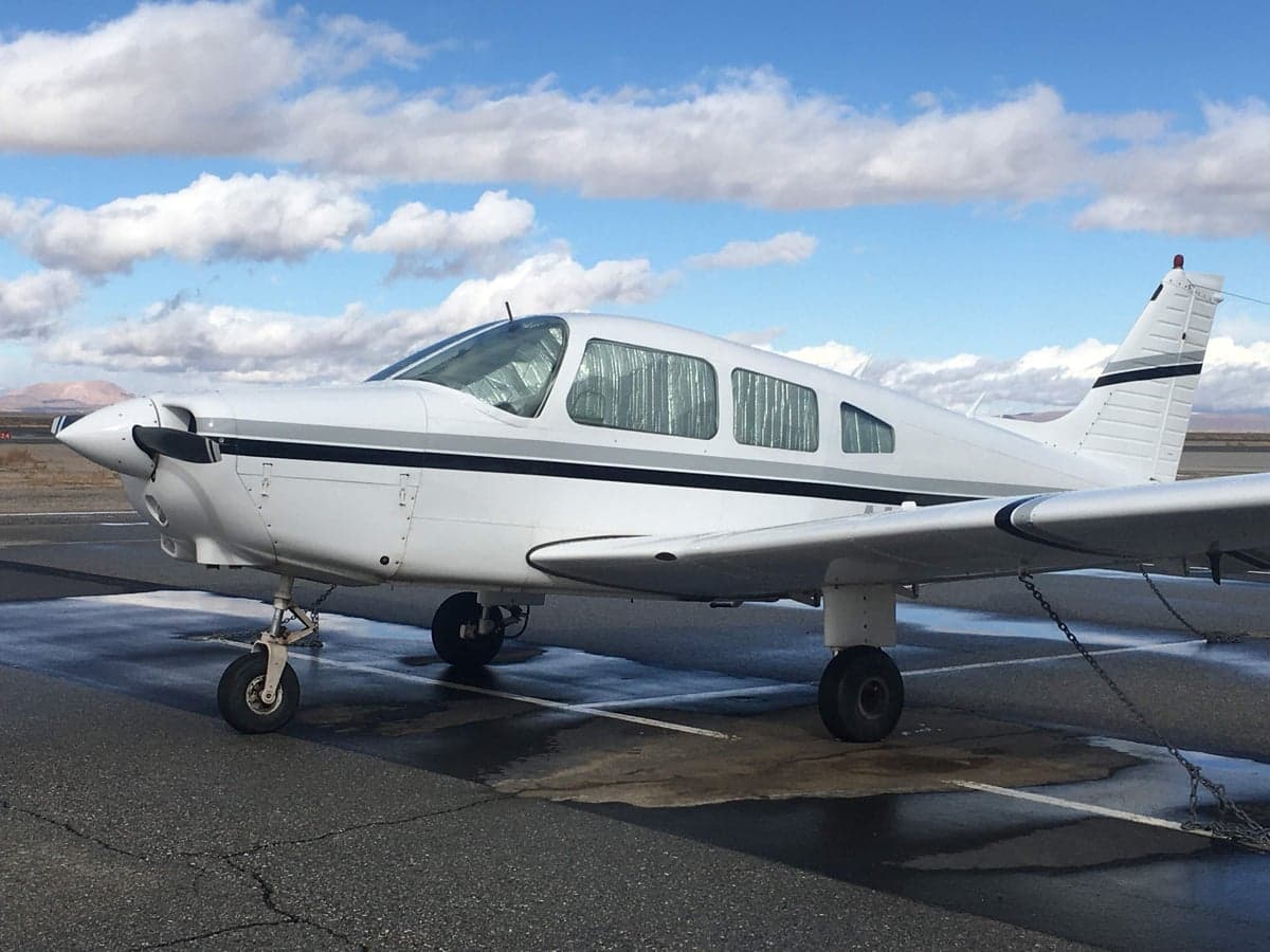A white Piper PA-28 Cherokee sits on the tarmac on a sunny day.