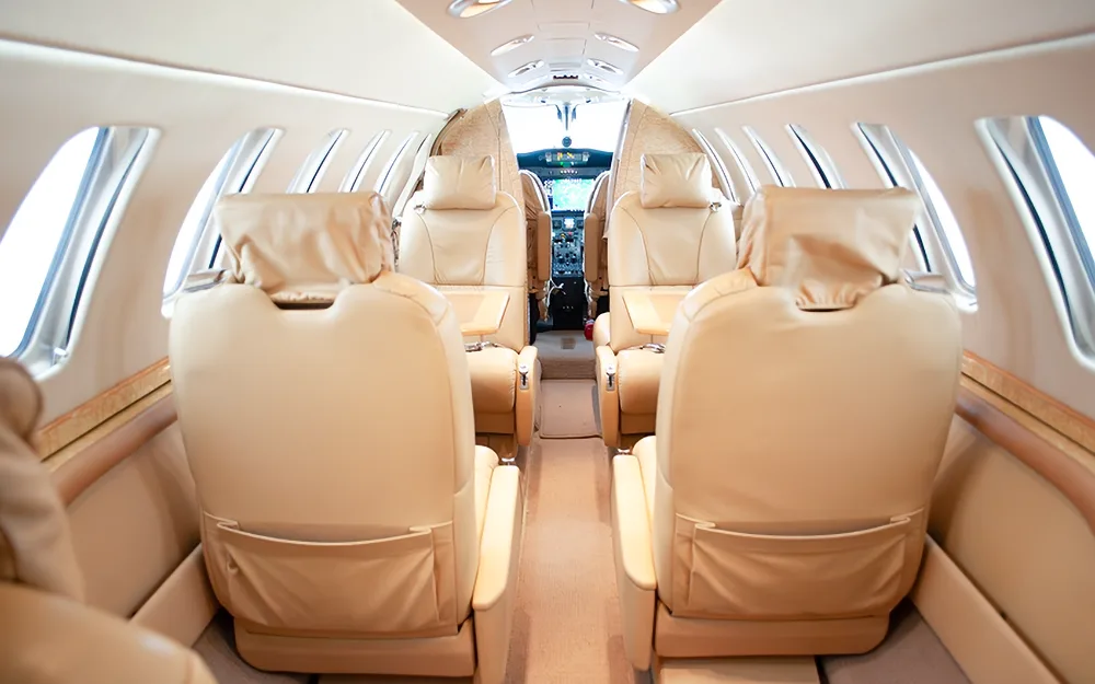 The interior of a business jet cabin, showing rows of beige leather seats leading towards the cockpit.