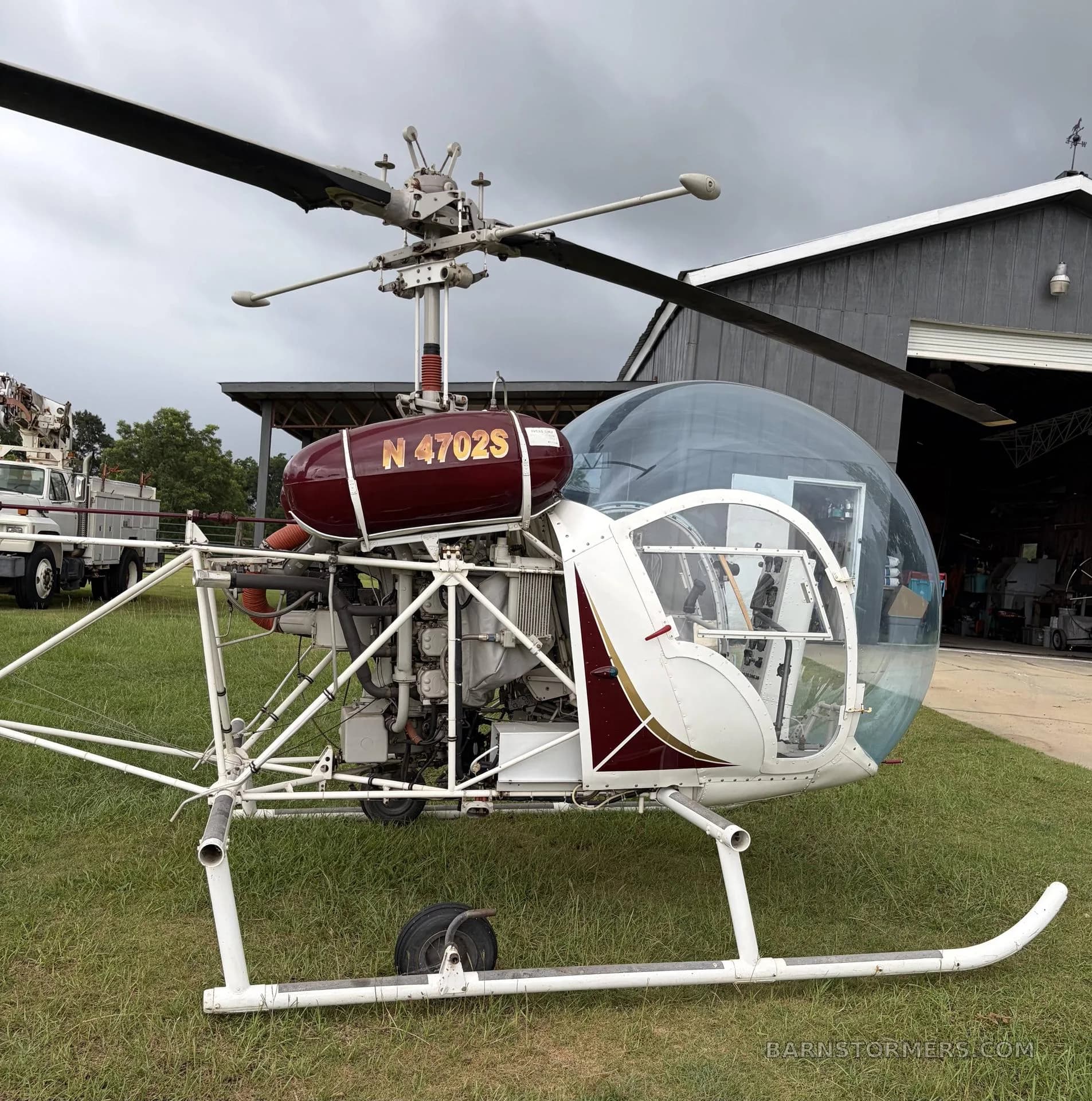A red and white helicopter with registration N4702S parked on grass in front of a hangar.