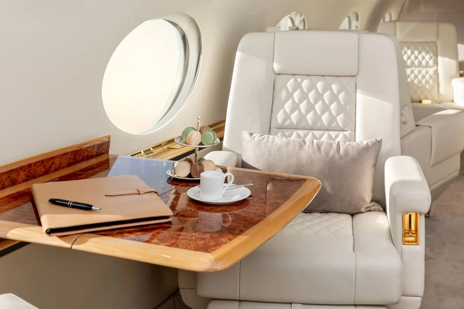 A luxurious aircraft interior with white leather seats, a wooden table, and a window.