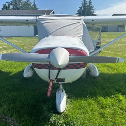 A Cessna 150 aircraft with a white and black paint scheme and red accents, covered with a gray cover.