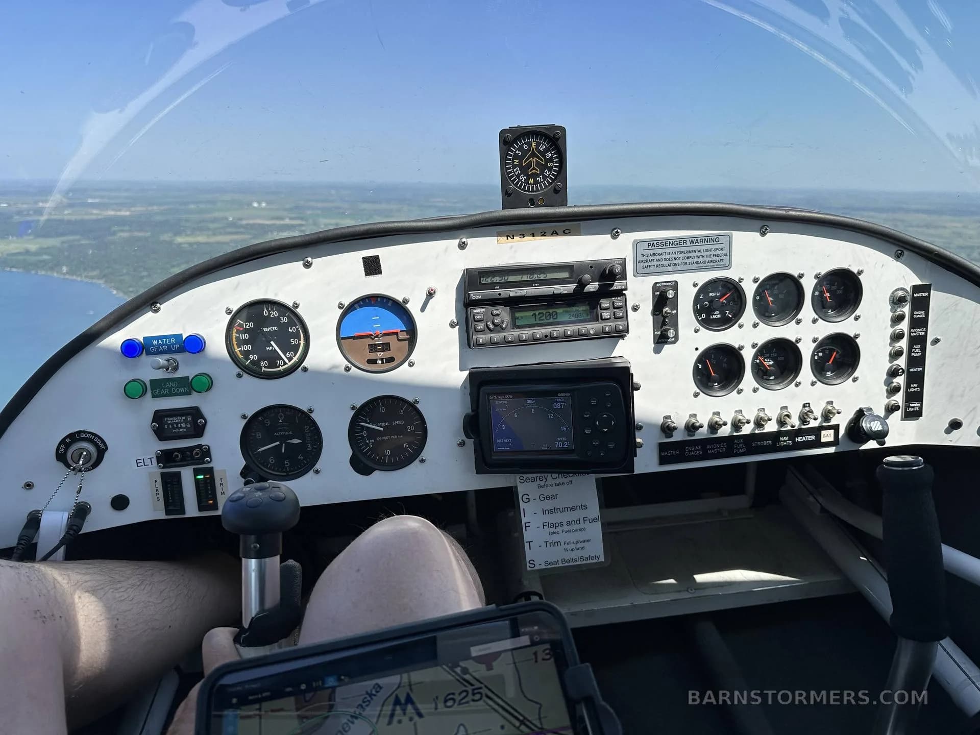 The cockpit of a Searey amphibian aircraft shows the instrument panel, controls, and a view of a lake and land from above.