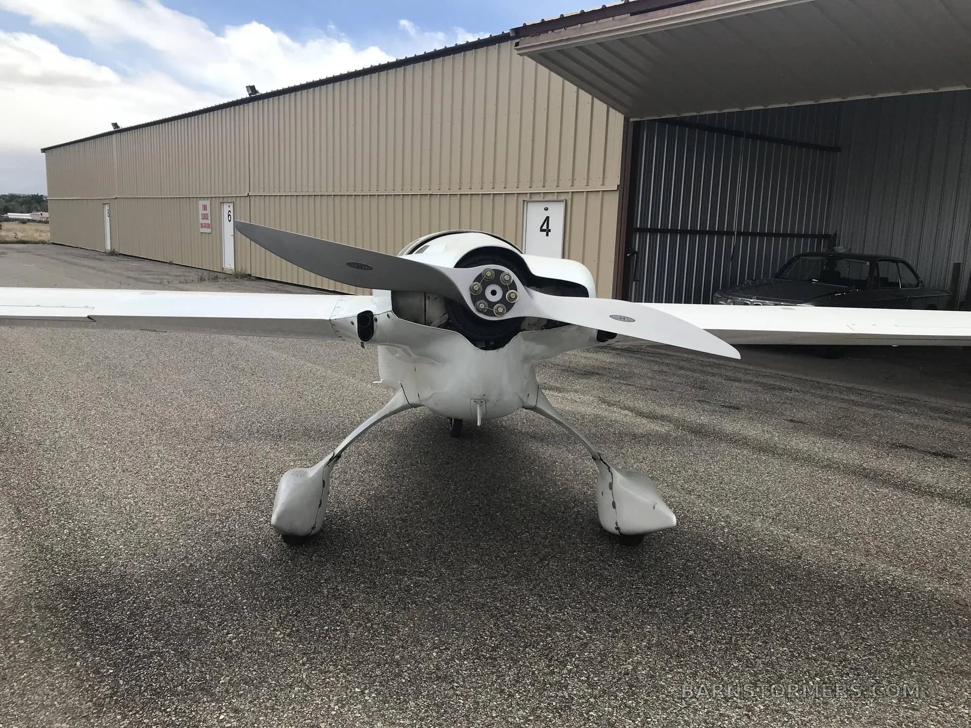 A white, propeller-driven aircraft with a canard wing is parked in front of a metal hangar, viewed from the front.