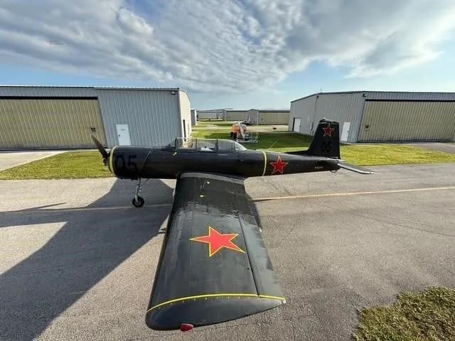 A dark gray Yak-52 aircraft with red stars and yellow accents, viewed from the rear-left, parked on an asphalt ramp.