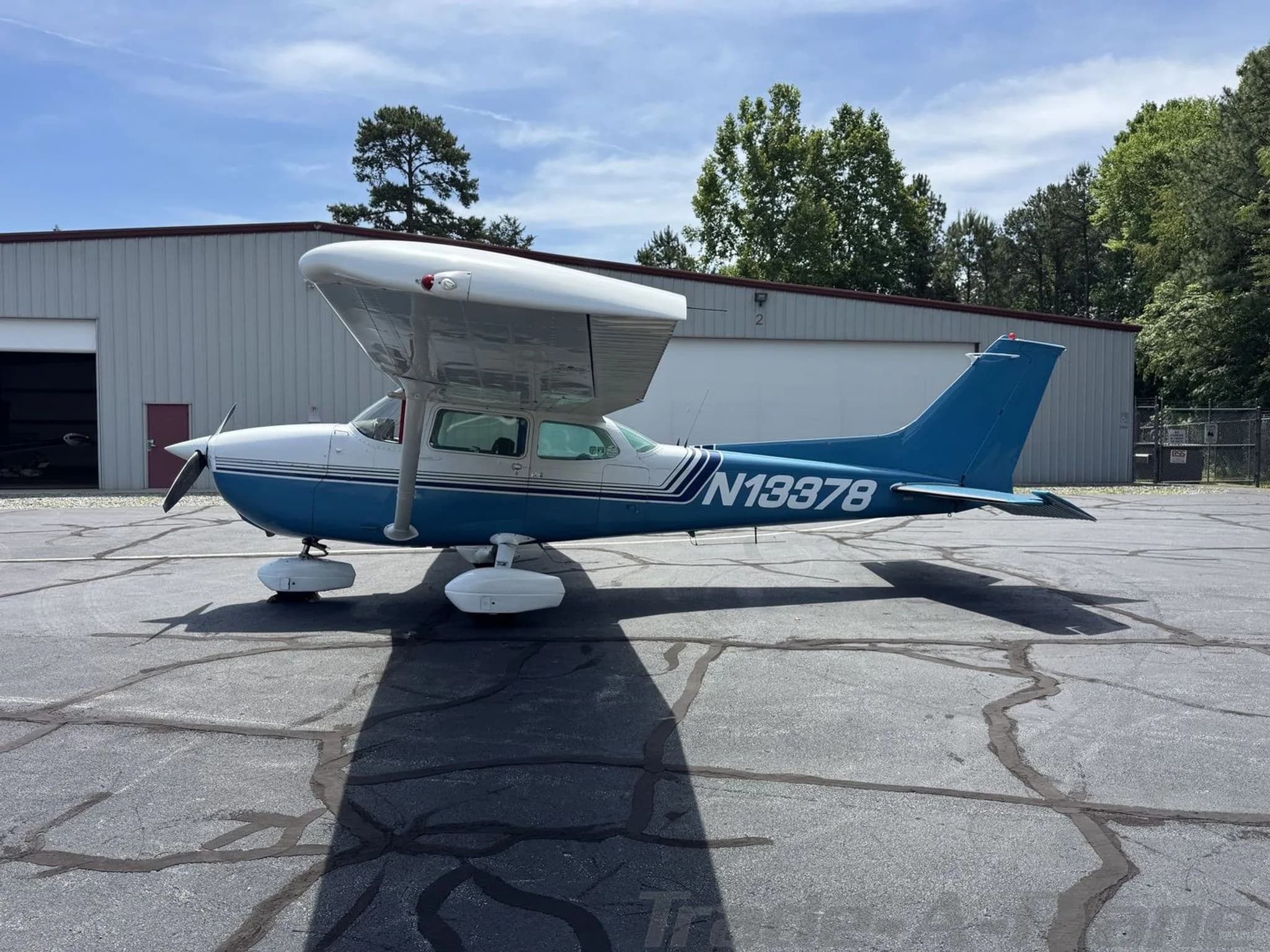 A blue and white Cessna 172 Skyhawk (N13378) parked on an asphalt apron next to a hangar.
