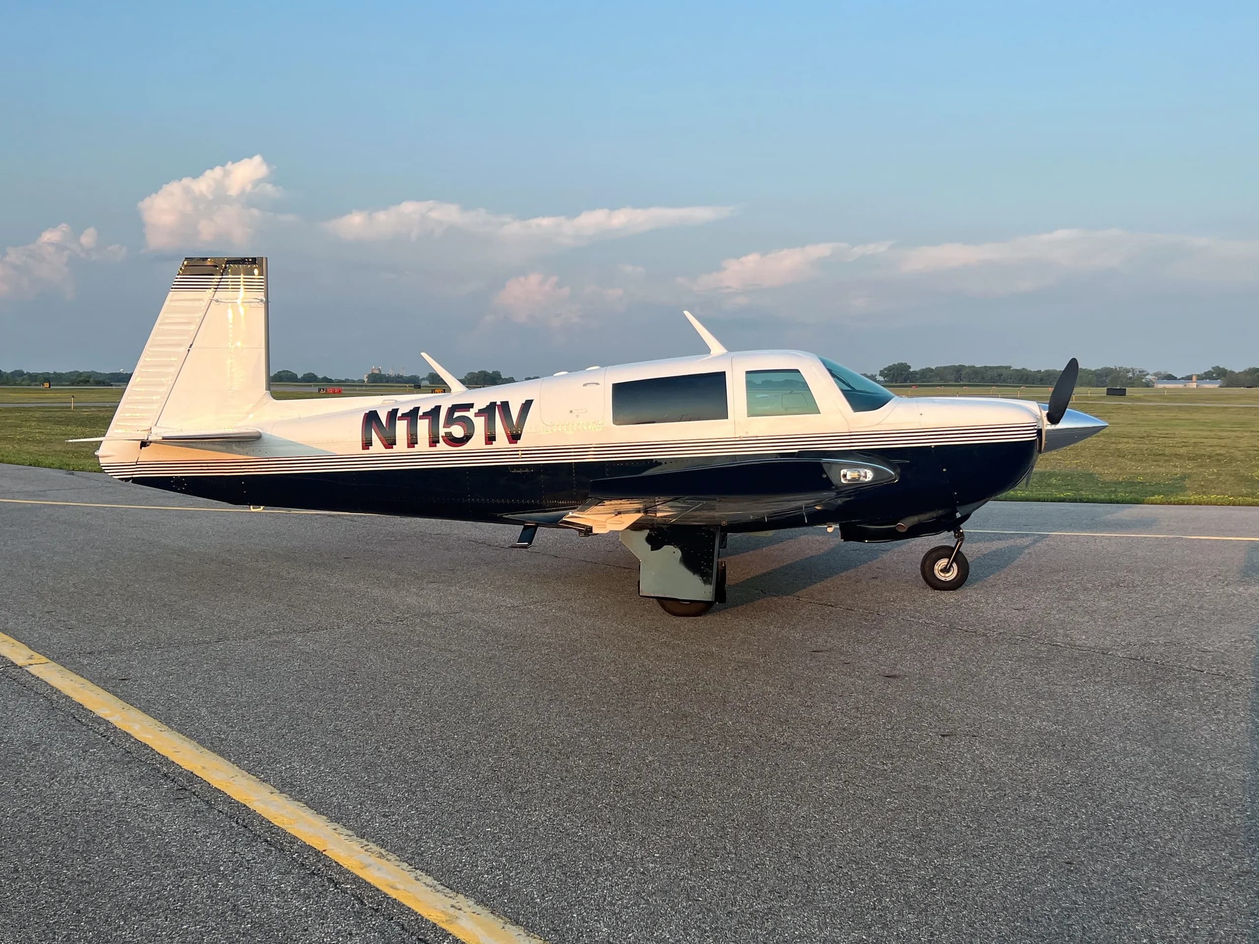 A white and dark blue single-engine aircraft, N1151V, parked on an asphalt runway with a cloudy sky.
