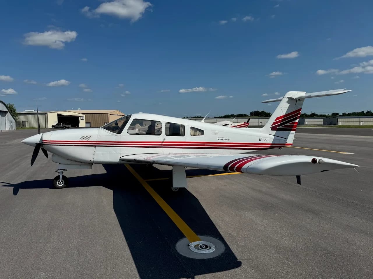 A white Piper Arrow II aircraft with red and brown stripes parked on an asphalt tarmac, viewed from the left side.