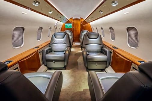 The luxurious interior of a business jet cabin with grey leather seats and wood paneling, viewed from the rear.