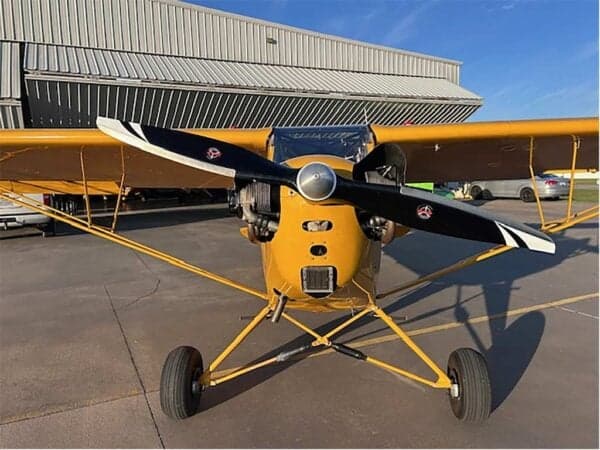 Front view of the yellow Legend Cub aircraft N90LC with its propeller, parked in front of a hangar.