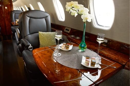 A close-up of a private jet's cabin table set with white wine, desserts, and a vase of white orchids next to a black leather seat.