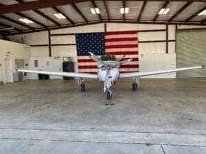 Front view of a Beechcraft Bonanza aircraft parked in a hangar with an American flag in the background.