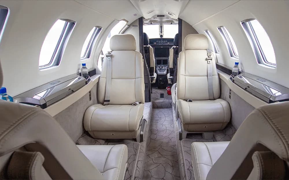 The aircraft cabin interior features light-colored leather seats, a patterned carpet, and a view towards the cockpit.