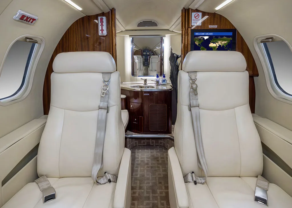 The rear section of a private jet cabin, with two light-colored seats facing a screen displaying a world map and a lavatory entrance.