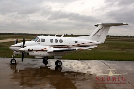 A white Beechcraft King Air twin-turboprop aircraft with dark red and gold stripes is parked on a wet tarmac under a cloudy sky.