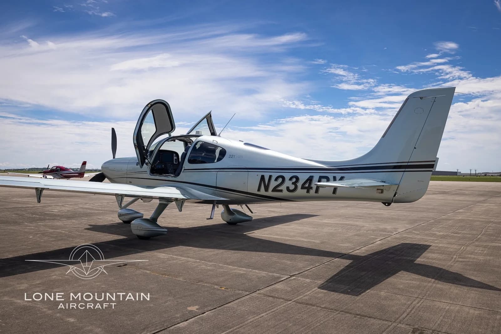 A silver Cirrus SR22 aircraft (N234PY) with open doors on a tarmac, with another red aircraft in the distance.