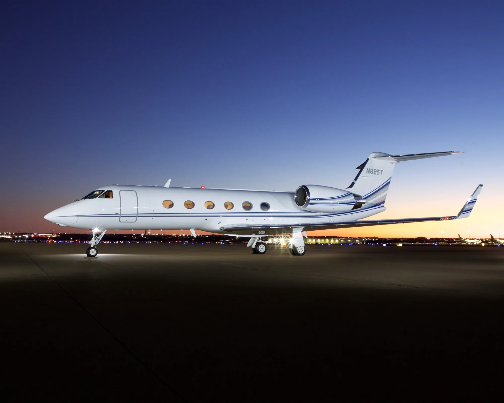 A white Gulfstream G550 aircraft with blue stripes, registration N825T, is parked on an airport tarmac at dusk.