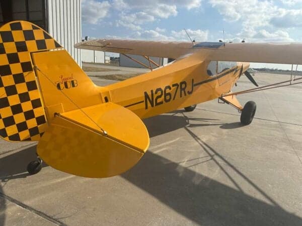 Rear three-quarter view of a yellow Legend Cub aircraft with registration N267RJ and a black and yellow checkered tail.