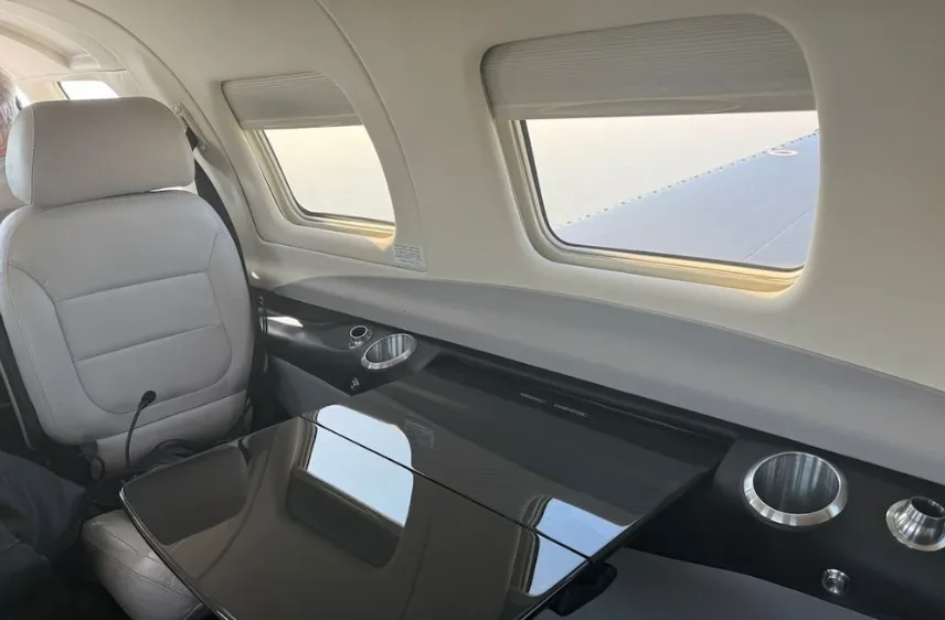 The aircraft's cabin interior features light grey seats, a dark table, and windows looking out at the wing.