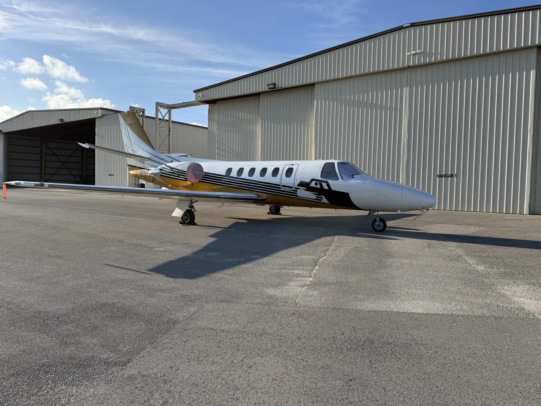A Cessna Citation jet, N917GP, with white and gold paint scheme, parked on an airfield next to a large hangar.