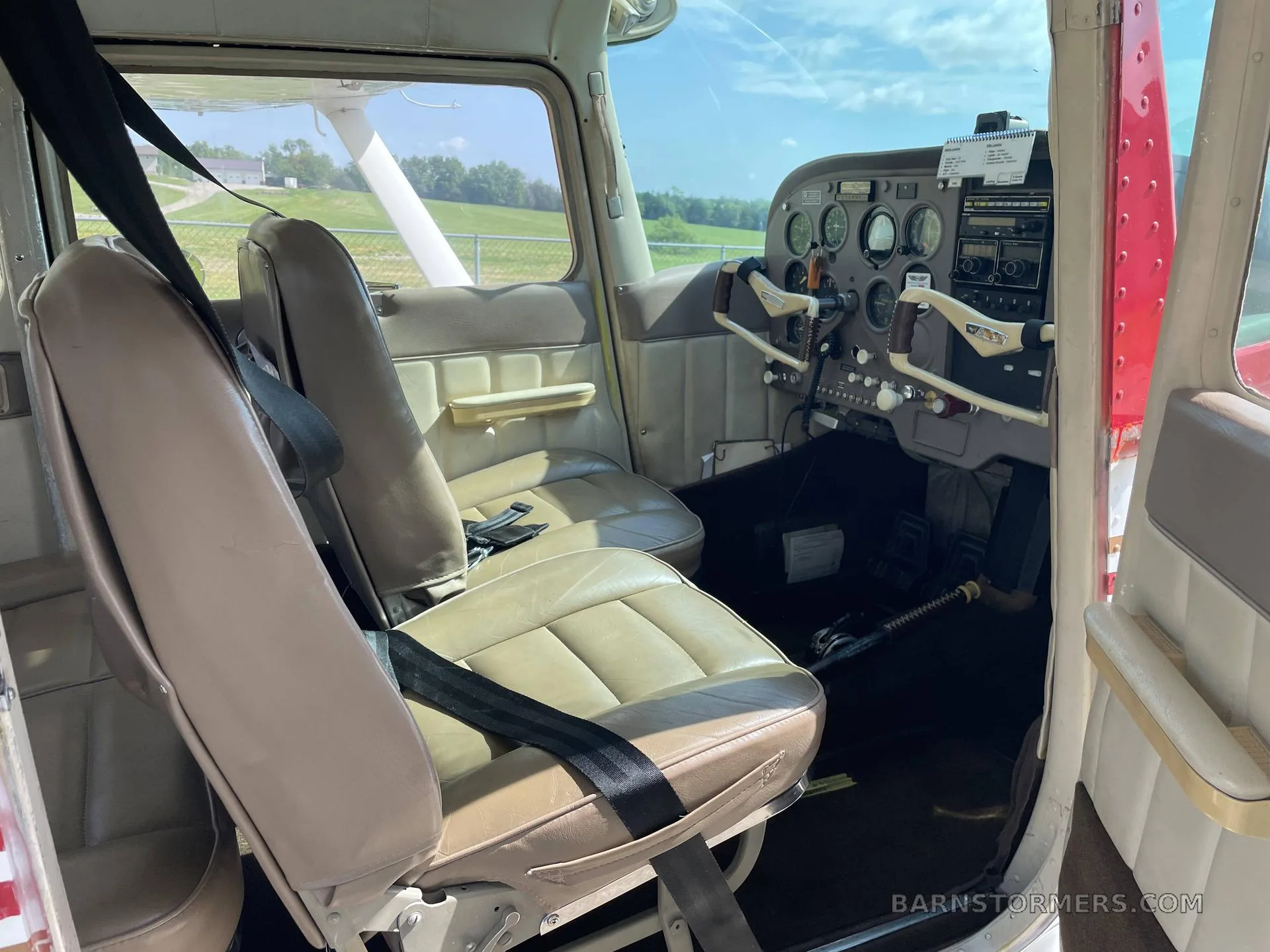 The interior of a Cessna aircraft, showing the front two tan seats with seatbelts and the cockpit beyond.