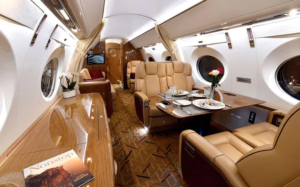 A private jet cabin interior featuring beige seats, a dining table set with plates, and a magazine on a side table.