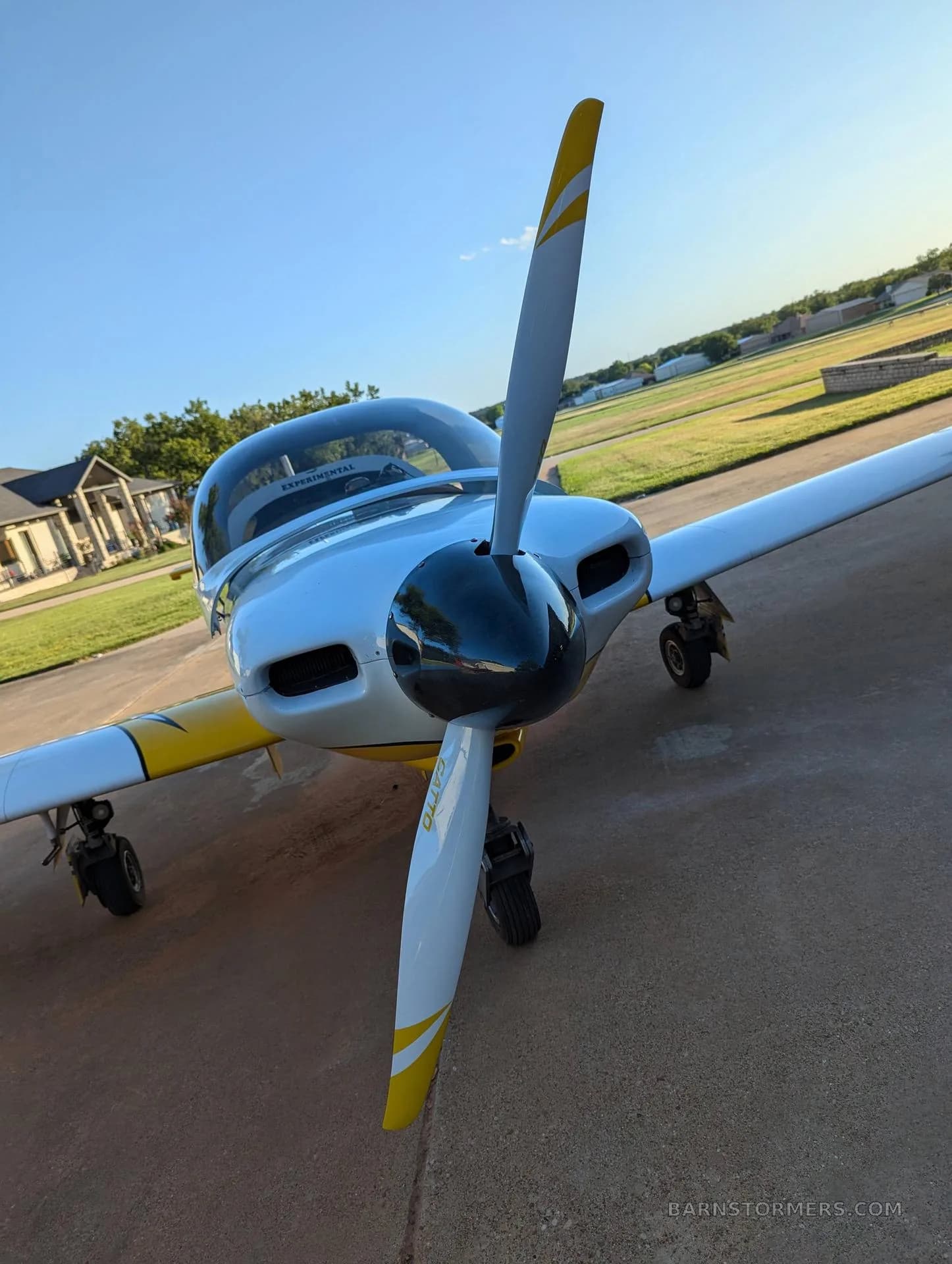 A close-up front view of a white and yellow single-engine aircraft with a black propeller spinner, parked on concrete.