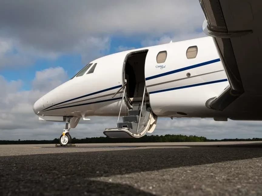 A close-up view of the front fuselage of a white Cessna Citation XLS+ business jet with blue stripes, parked on a tarmac with its main door open.