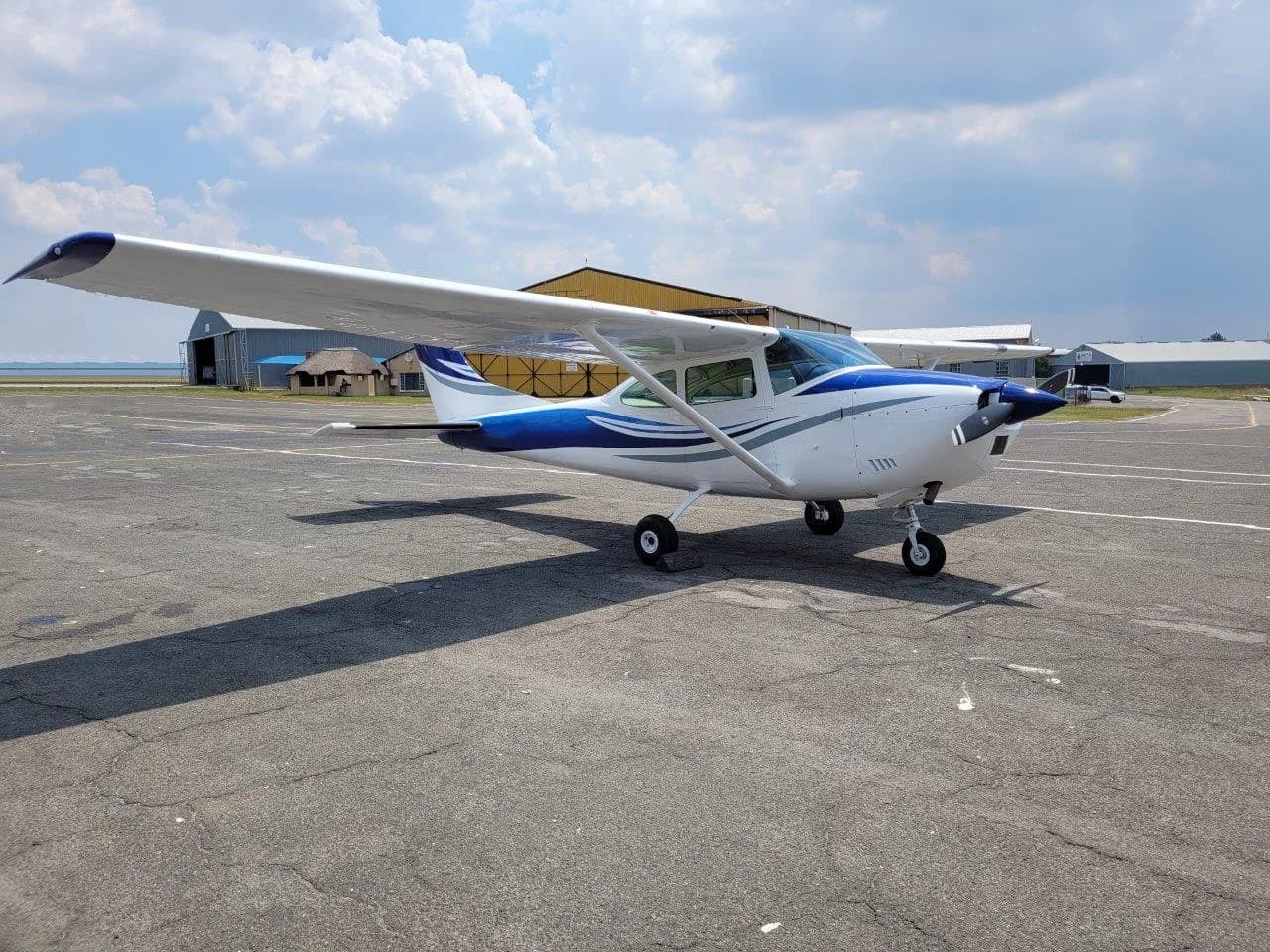 A white and blue Cessna 182 Skylane aircraft is parked on an asphalt tarmac under a partly cloudy sky.