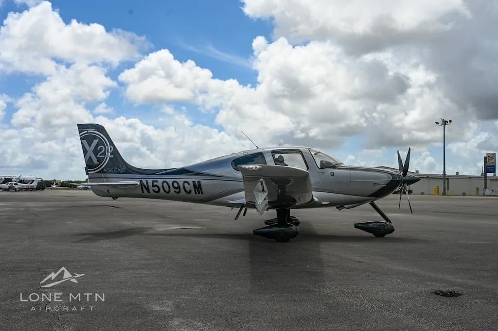 A silver and black Cirrus SR22 aircraft with registration N509CM parked on an airfield, viewed from the left side with buildings in the background.