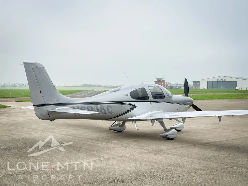 A silver Cirrus SR22 aircraft, registration N6918C, parked on an airfield with a control tower in the distance.