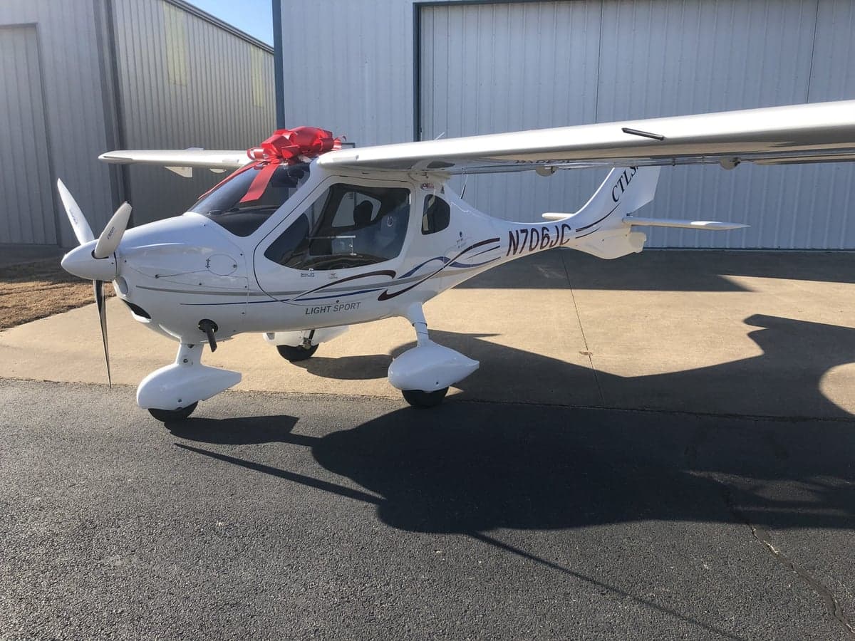 A white CTLS light sport aircraft with a red bow on the nose is parked on the tarmac.