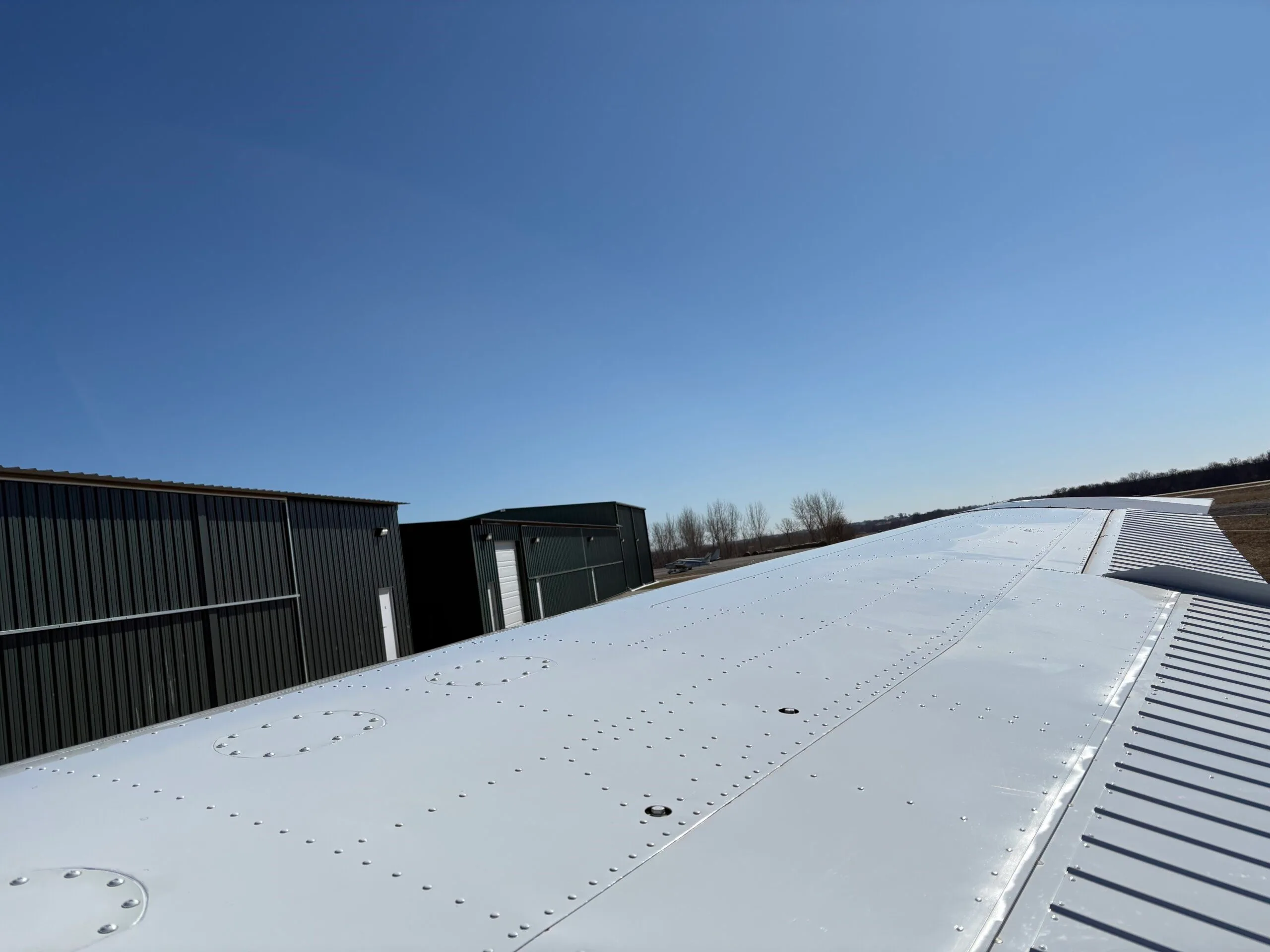 View along the top of the white wing of an aircraft, with hangars in the background.