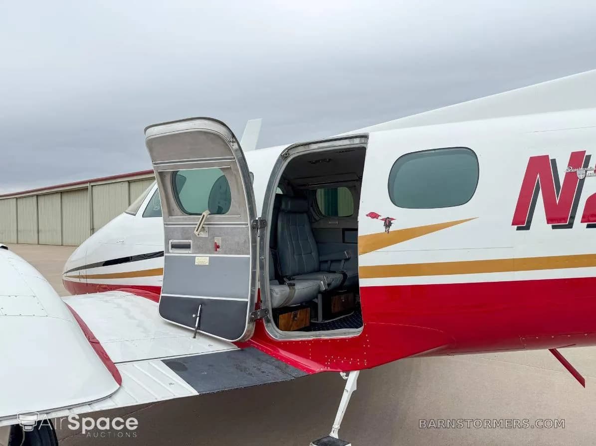 The side of a white and red twin-engine aircraft with gold accents, showing the open passenger door and a glimpse of the cabin interior.