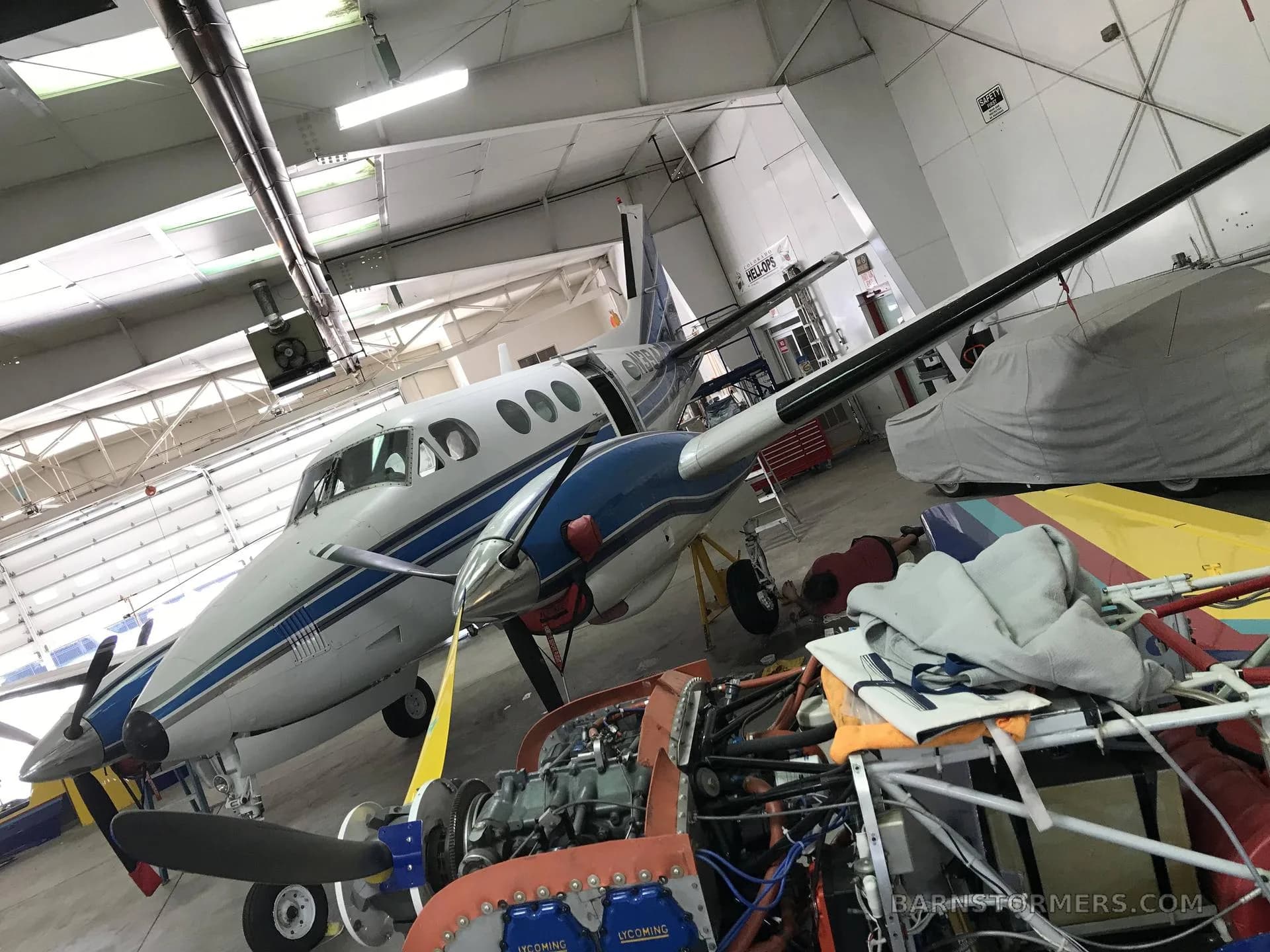 A white and blue twin-engine turboprop aircraft is parked in a hangar, with an open-engine aircraft frame in the foreground.