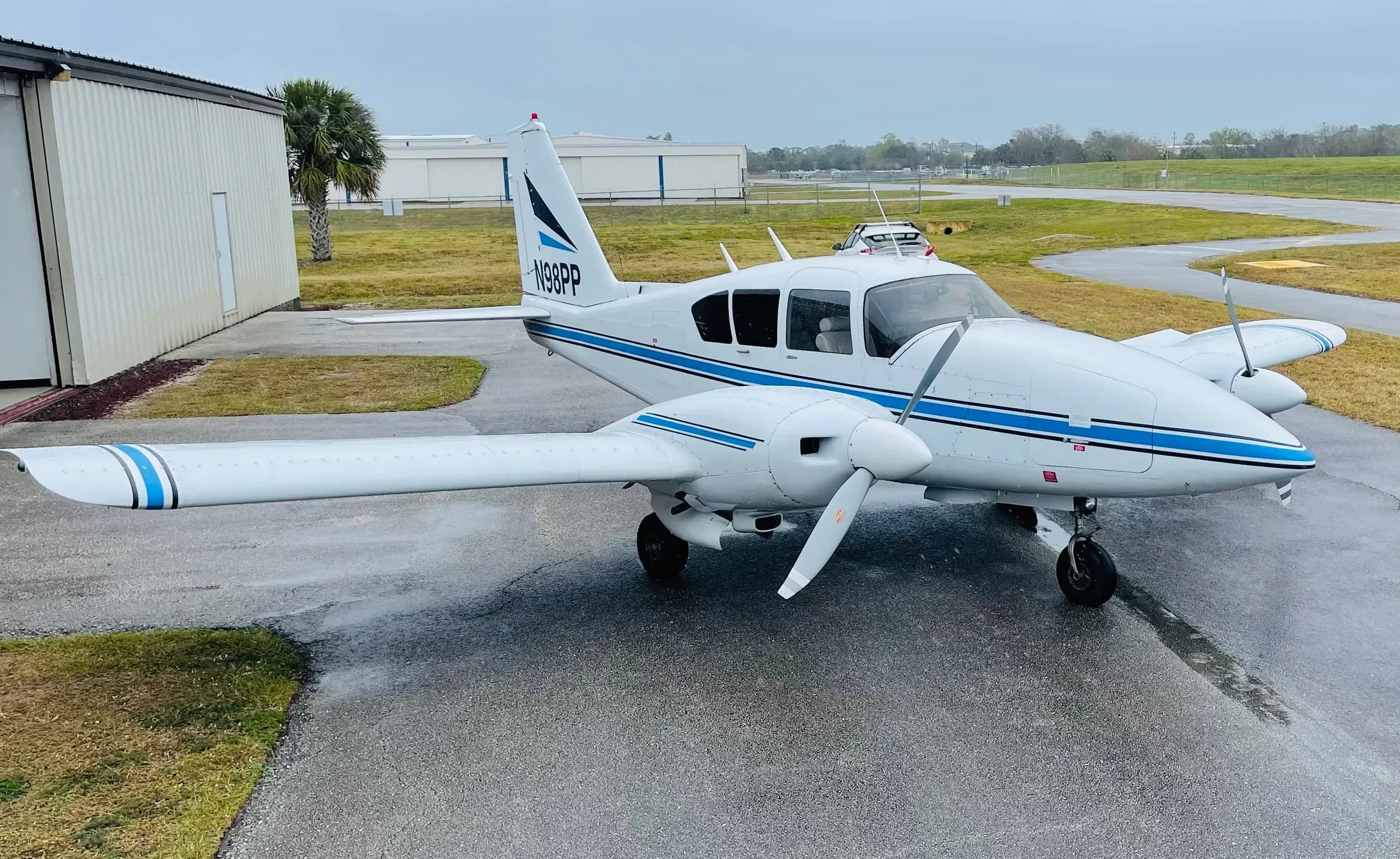 A white Piper Aztec aircraft with blue stripes parked on a wet tarmac with a building and palm trees in the background.