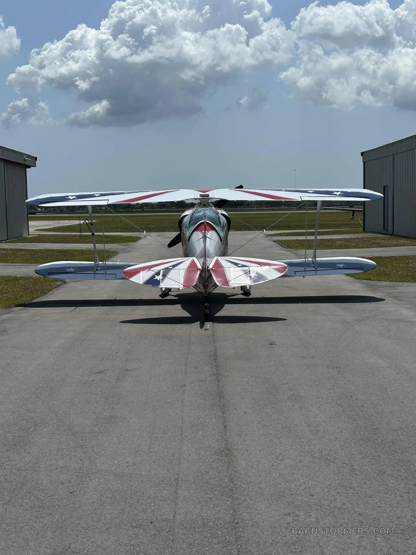 A Pitts Special biplane with a red, white, and blue patriotic paint scheme on the tail and lower wings, viewed from the rear on an airfield.