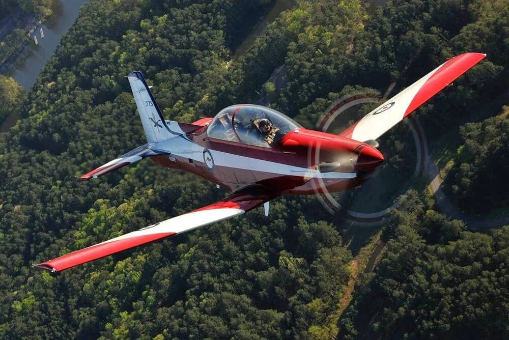 A red and white single-engine aircraft with a bubble canopy flies over a dense green forest.