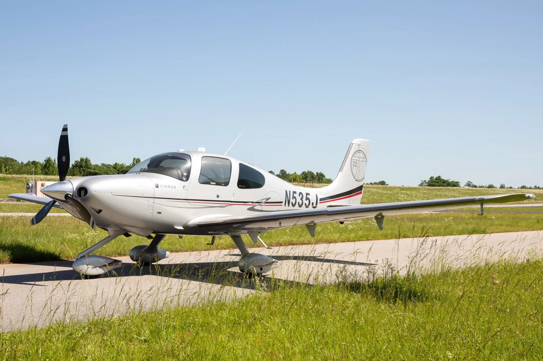 A silver Cirrus SR22T aircraft with registration N535J is parked on an asphalt strip next to green grass under a clear blue sky.