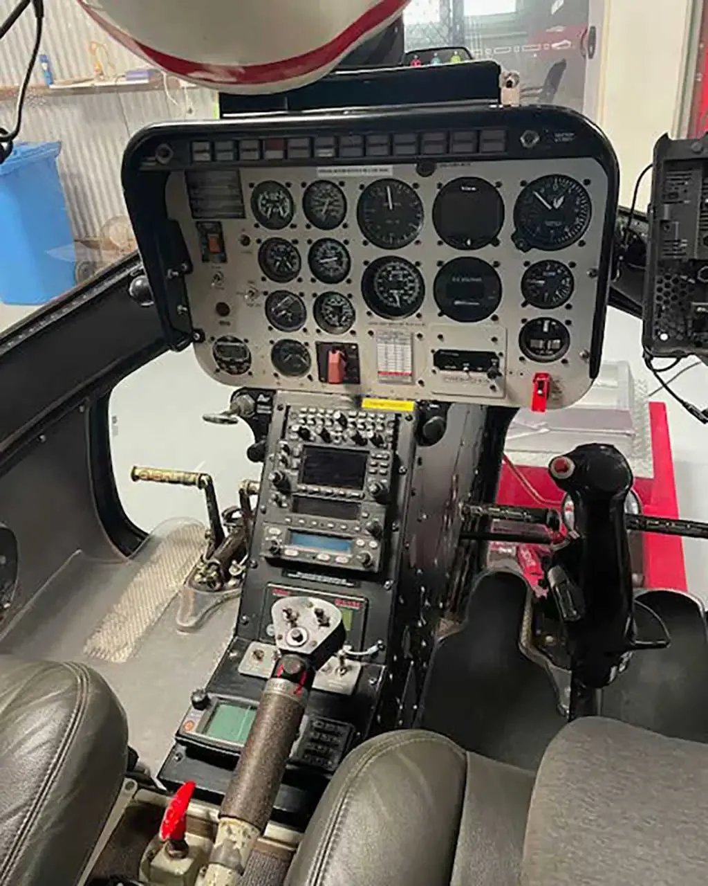 An interior view of a helicopter cockpit, showing the instrument panel, cyclic, collective, and pilot's seat.