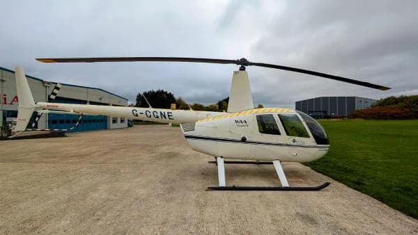A white Robinson R44 Raven II helicopter with registration G-CGNE and yellow tail stripes parked on an airfield.