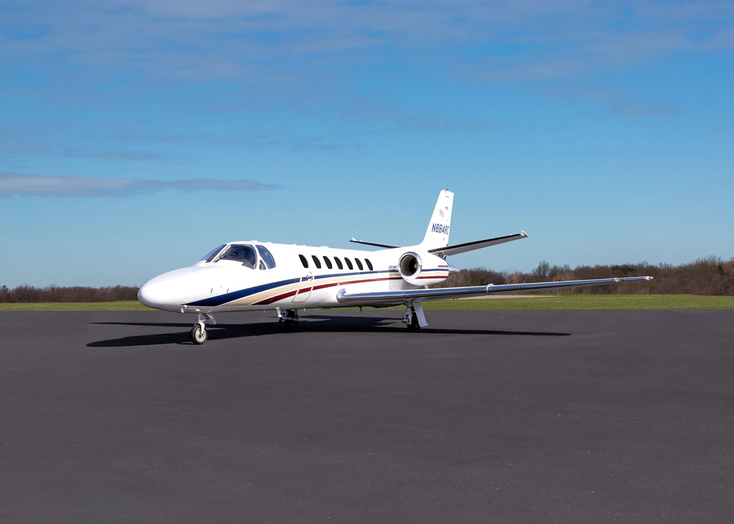 A white business jet with blue and red stripes, registration N884RC, parked on an asphalt tarmac under a blue sky.
